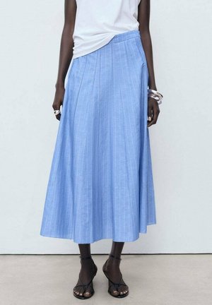 Femme portant une jupe midi bleu clair à fines rayures, associée à un haut blanc et des sandales noires à brides, debout contre un mur blanc uni.