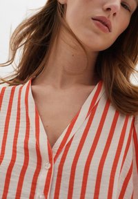 Chemise à boutonner en tissu crème avec de larges rayures verticales rouges, présentant un décolleté en V et une finition texturée.