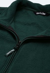 Grüne Fleecejacke mit hohem Kragen und Reißverschluss, ausgestattet mit einer strukturierten Oberfläche und einem Etikett mit der Aufschrift "switcher."