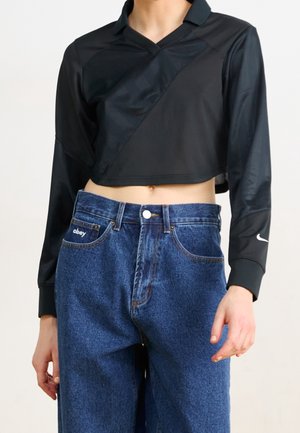 Personne portant un crop top noir à manches longues avec un col et un jean bleu taille haute avec "obey" sur la poche avant.