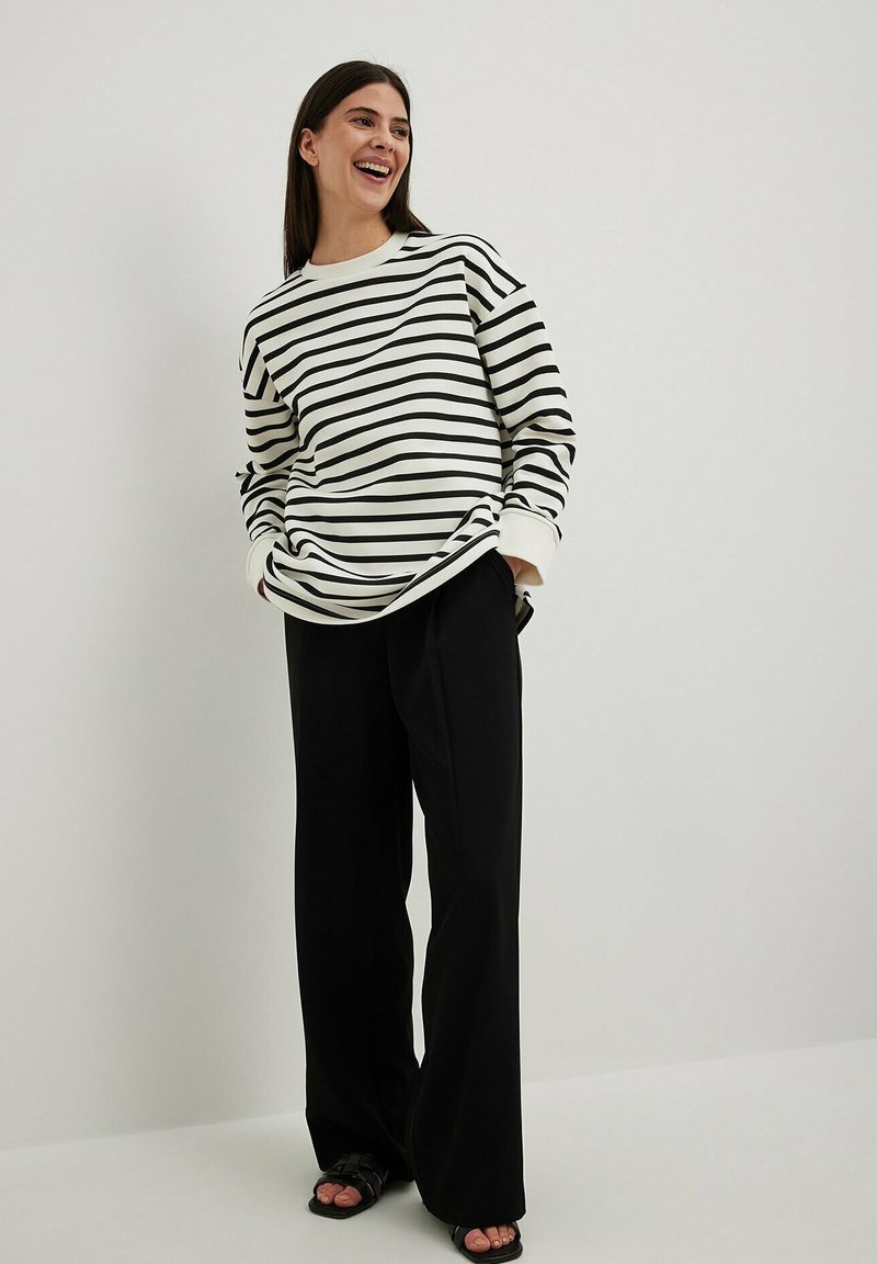Chemise à manches longues rayée blanche et noire avec une coupe décontractée, associée à un pantalon noir ample et des sandales noires, sur un fond uni.
