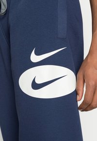 Primo piano di una mano di una persona che riposa su pantaloni tuta blu navy con grandi loghi bianchi del marchio Nike sulla coscia.
