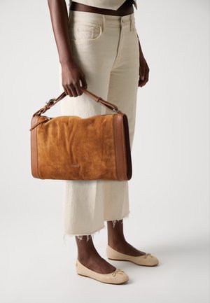 Brown suede handbag with leather accents, zip closure, and structured shape, held in hand. Worn with light-colored cropped trousers and flats.