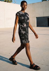 Kvinde går udendørs iført en sort og grå tie-dye ærmeløs kjole og sorte platformsandaler i klart sollys.