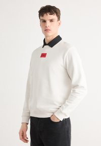 Kerge beež crewneck-sviiter musta kaelusega, millel on punane ristkülikukujuline logo-patch rinna peal. Sile tekstuur, juhuslik disain.