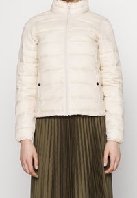 Jasnokremowa pikowana kurtka z suwakiem z przodu i kieszeniami na zatrzask, noszona na oliwkowej plisowanej spódnicy.