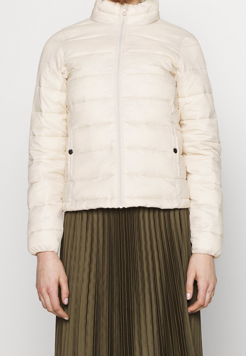 Jasnokremowa pikowana kurtka z suwakiem z przodu i kieszeniami na zatrzask, noszona na oliwkowej plisowanej spódnicy.