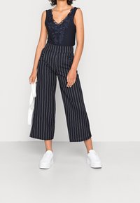 Granatowy bodysuit z koronkowymi detalami w kwiaty, w zestawieniu z czarno-białymi spodniami o szerokich nogawkach oraz białymi sneakersami. Modelka trzyma białą torbę.