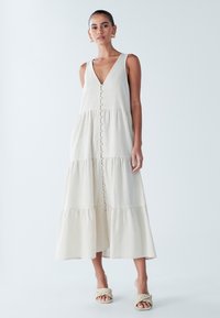 Beżowa, bezrękawnikowa sukienka z głębokim dekoltem w kształcie V, z przodu na guziki i o falbaniastym zakończeniu. Wykonana z lekkiego materiału, z subtelną fakturą.