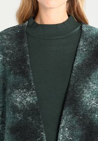 Cardigan verde, texturizzato, con una sfumatura che va dal chiaro allo scuro, apertura frontale, abbinato a un top fitted verde scuro a collo alto.