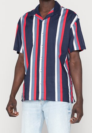 Homme portant une chemise à manches courtes boutonnée avec des rayures verticales bleu marine, rouge, blanc et bleu clair, associée à un jean bleu clair.