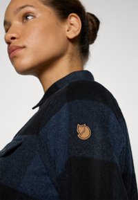 Patches flanellen overhemd in donkerblauw en zwart, met een schouderpatch van een voslogo. De stof heeft een gestructureerde, geweven uitstraling.
