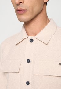 Beige shirt met structuur en een kraag, met donkere knopen en twee voorkanten zakken. De stof heeft een zachte, geweven uitstraling.