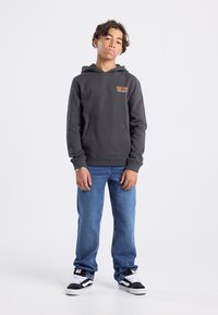 Sudadera gris oscuro con bolsillo canguro, logo en naranja en el pecho, combinada con jeans de mezclilla azules y zapatillas en blanco y negro.