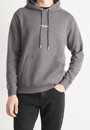 Homme portant un sweat à capuche gris avec une poche avant et un pantalon noir, main droite dans la poche, bras gauche détendu le long du corps.