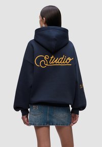 Felpa navale con ricamo giallo "Studio" sul retro, taglio oversized e cappuccio con cordoncino. Abbinata a una minigonna di jeans.