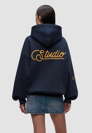 Marineblauer Hoodie mit gelber "Studio"-Stickerei auf dem Rücken, oversize Schnitt und Kapuze mit Kordelzug. Kombiniert mit einem Jeans-Minirock.
