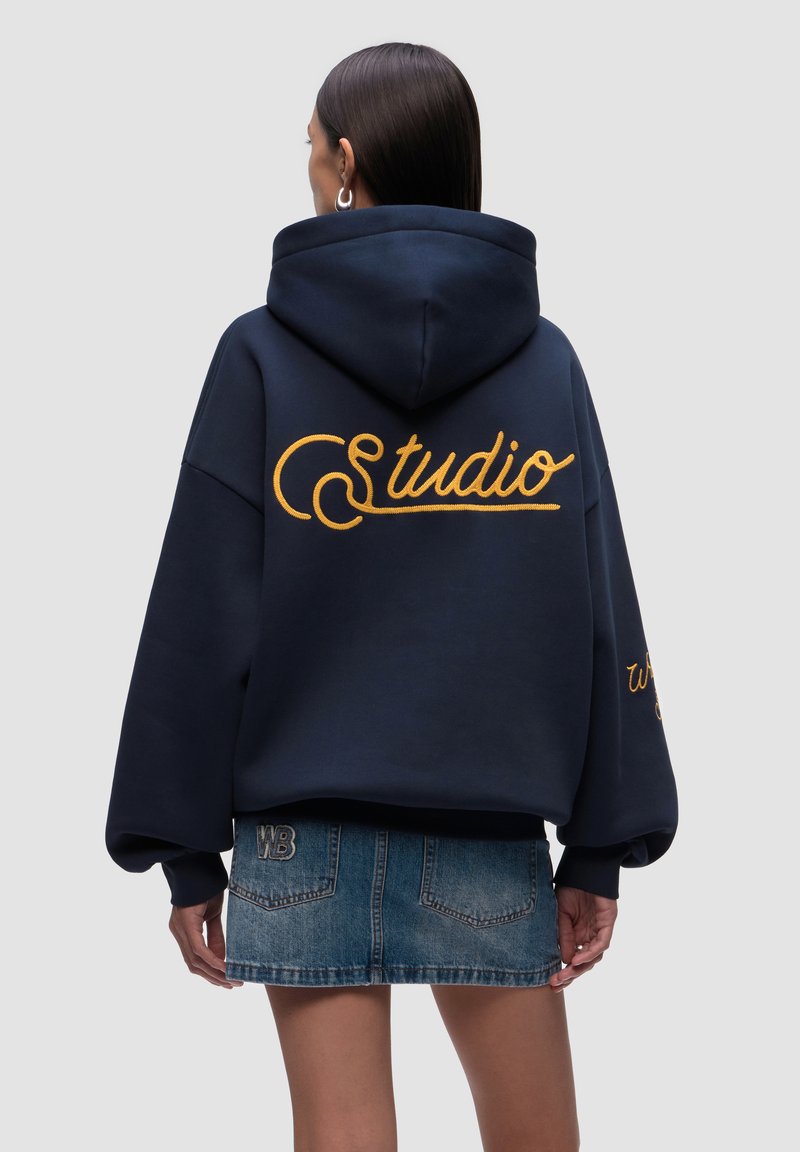 Felpa navale con ricamo giallo "Studio" sul retro, taglio oversized e cappuccio con cordoncino. Abbinata a una minigonna di jeans.