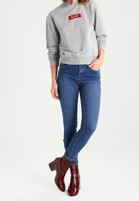 Sudadera gris con parche bordado rojo "NAKD", jeans ajustados azules y botas de tobillo burdeos con tacón grueso. Estilo básico y casual.