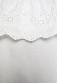 Weißer bestickter Stoff mit durchbrochenen Details und floralen Mustern entlang eines gewellten Randes, auf einem glatten, einfarbigen weißen Hintergrund.