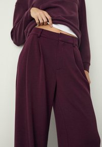 Burgunderfarbene, hochgeschnittene Hose aus weichem Stoff, mit weitem Bein, vorderen Falten und seitlichen Taschen. Dazu ein weißes Oberteil darunter.