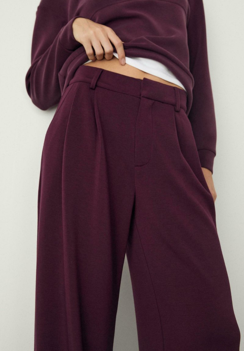 Burgunderfarbene, hochgeschnittene Hose aus weichem Stoff, mit weitem Bein, vorderen Falten und seitlichen Taschen. Dazu ein weißes Oberteil darunter.