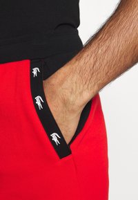 Röda bomullsjoggingbyxor med svart elastisk midjeband som har en vit logotyp. Ficka synlig, med kontrasterande svart kant.