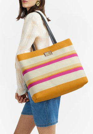 Donna con camicetta bianca e pantaloncini di jeans porta una grande borsa a righe con strisce orizzontali arancioni, rosa e beige e manici neri.