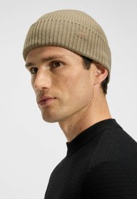 Gorro de punto acanalado beige con una pequeña etiqueta de logo. Llevado por una persona con pelo corto, vestida con un suéter negro texturizado, mirando ligeramente hacia la derecha.