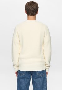 Kremfarvet strikket sweater med ribbet struktur, raglanærmer og rund hals. Kombineret med blå denimjeans med baglommer.