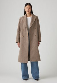 Dvojřadý kabát z texturované hnědé látky s klopovými reverzy, s dvěma velkými knoflíky, nošený přes bíl ý top a modré rozšířené džíny.