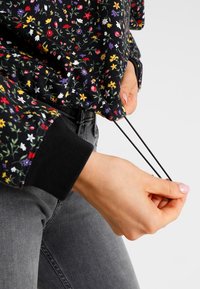 Sudadera negra con estampado floral, confeccionada en un tejido texturizado, que presenta patrones de flores multicolores y puños acanalados negros. Cuerdas visibles en el dobladillo.