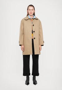 Cappotto impermeabile beige con un ampio colletto, chiusura frontale con bottoni e tasche laterali, indossato sopra un maglione a fantasia e pantaloni neri alla caviglia.