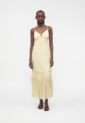 Femme portant une robe maxi sans manches jaune pâle avec des détails en dentelle à l'ourlet, debout devant un fond clair uni.