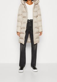 Abrigo acolchado beige con capucha, con un diseño acolchado y cremallera frontal. Lleva una camisa blanca debajo, combinado con jeans negros de pierna recta y zapatos negros.