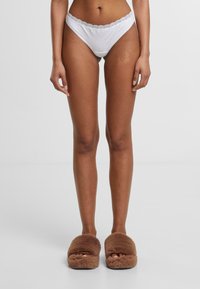 Witte katoenen bikini met kanten afwerking, gedragen met zachte bruin fluffy slippers. De bikini heeft een low-rise snit en een verfijnd ontwerp.
