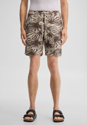 Mann trägt beige-braune Shorts mit Blumenmuster und Kordelzug in der Taille, weißes Hemd und schwarze Sandalen mit Schnalle, steht vor einem einfarbigen Hintergrund.