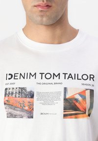 Camiseta blanca de algodón con cuello redondo, que presenta un texto en negrita de color negro y estampados gráficos coloridos en la parte frontal. Marca: Denim Tom Tailor.