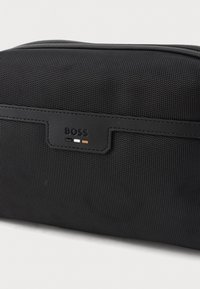 Svart strukturert reiseveske med et lite lærmerke preget med merket "BOSS" og tre små fargeblokker under.