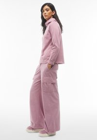 Felpa con cappuccio rosa chiaro a maniche lunghe abbinata a pantaloni cargo larghi con tasche laterali. Il modello indossa sneakers chiare.