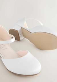 Pantofi cu toc alb, cu un exterior sintetic neted, vârf deschis, curea reglabilă la glezna și toc bloc scăzut, cu o talpă texturată.