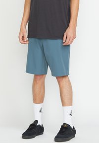 Shorts teal en tissu léger, associés à un t-shirt gris foncé, des chaussettes blanches avec logo et des baskets noires au design profil bas.