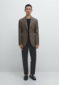 Bruine geweven blazer met notched revers, gecombineerd met donkere broeken en zwarte loafers. Slim fit, gestructureerde stof en sluiting met twee knopen.