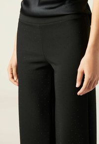 Zwarte wijde broek van gladde stof, met een subtiel stippenpatroon en kleine zilveren accenten, en een op maat gemaakte tailleband.