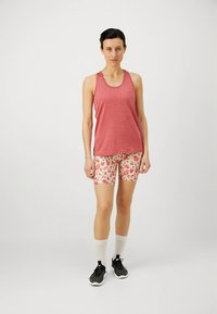 Rotes ärmelloses Tanktop kombiniert mit hellen Shorts mit Blumenmuster, dazu weiße Socken und schwarze Sportschuhe.