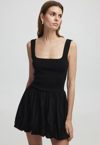 Robe noire sans manches avec un corsage ajusté en maille côtelée et une jupe évasée en tissu texturé. Décolleté carré et détail de ceinture froncée.
