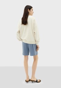 Beige oversized sweatshirt, blauwe shorts en zwartbruine haksandalen. Glad textuur, ontspannen pasvorm, met zichtbare stiksels op de kleding.