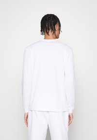 T-shirt blanc à manches longues présentant un col rond, une coupe décontractée et un matériau en coton doux. Les coutures sont minimales, sans logos visibles.