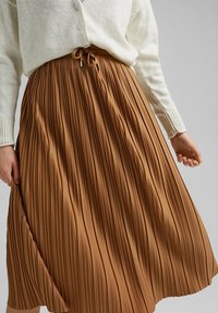 Femme portant un cardigan beige et une jupe plissée marron mi-longue avec une taille à cordon, debout devant un fond gris uni.