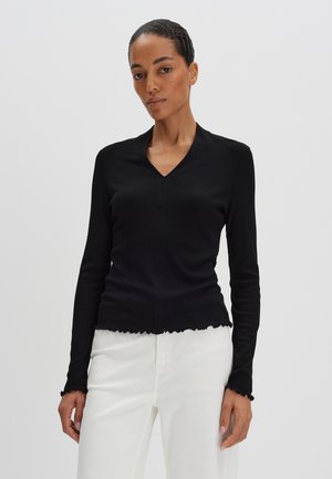 Top noir à manches longues avec un décolleté en V, texture côtelée et ourlets et poignets à volants. Porté avec un pantalon blanc pour le contraste.